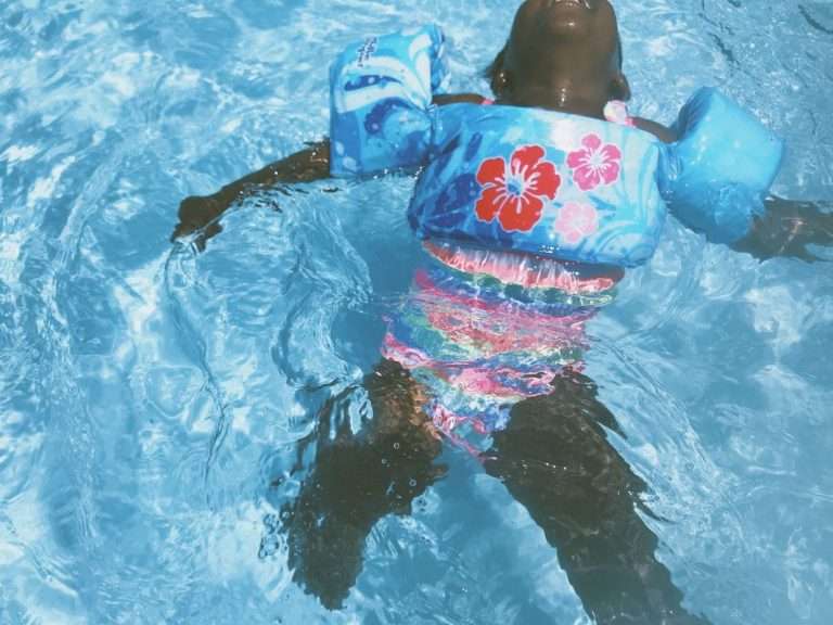A child wearing a floral swimsuit and arm floaties floats on their back in a sunlit swimming pool.
