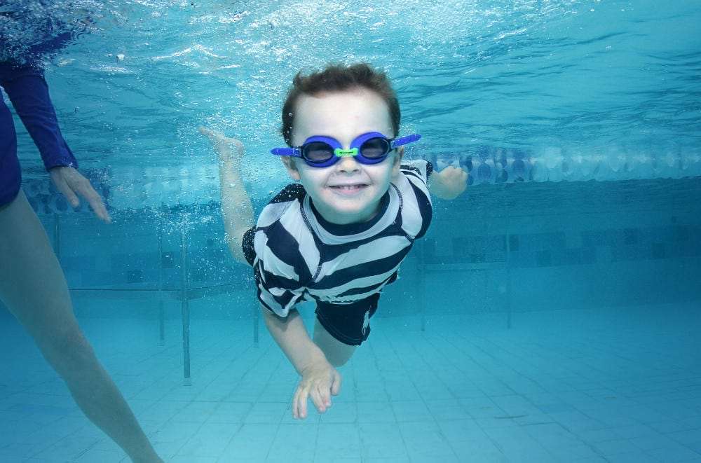 Young boy with goggles joyfully swimming underwater in a pool, showcasing his super swim skills, with an adult partially visible to the side.