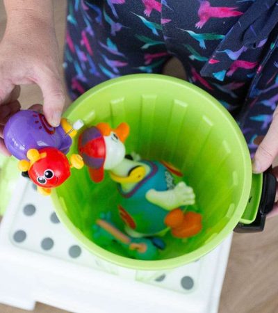 An overhead view of a person holding a purple toy over a green bucket filled with various colorful children's super therapy toys.